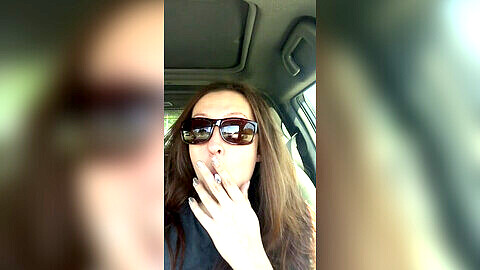Smoking goddess D complains about the line while puffing on a cigarette and wearing sunglasses in the car