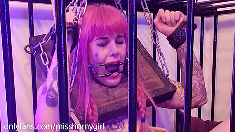 Jeune fille soumise enchaînée dans une cage et un carcan, baisée par une machine impitoyable